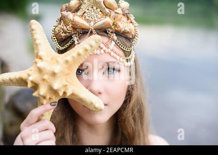 Fantasy Frau echte Meerjungfrau mit Dreizack Mythos Göttin des Meeres mit goldenen Schwanz sitzt in Sonnenuntergang auf Felsen.. Gold Haar Krone Muscheln Perlen Schmuck. Merma Stockfoto