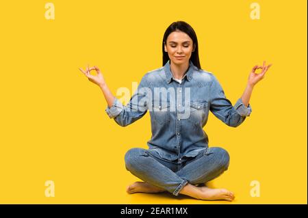 Ruhe bewahren. Foto einer ziemlich ruhigen kaukasischen jungen Frau mit geschlossenen Augen, meditiert in Lotusposition auf isoliertem gelben Hintergrund Stockfoto
