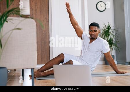 Afroamerikanischer Mann, der Training mit Online-Trainer mit Laptop zu Hause im Seitenbrett stehend macht. Stockfoto