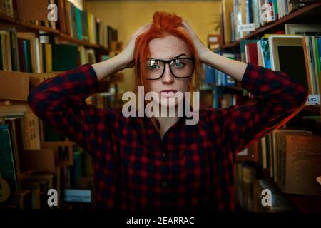 Ein Mädchen mit roten Haaren steht in der Bibliothek, trägt eine Brille, hat rote Haare und hält den Kopf Stockfoto