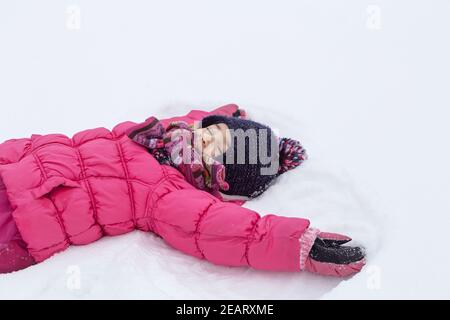 Ein kleines Mädchen in einer rosa Jacke macht einen Engel auf dem frisch gefallenen Schnee. Winter Kinder Spaß Konzept. Stockfoto