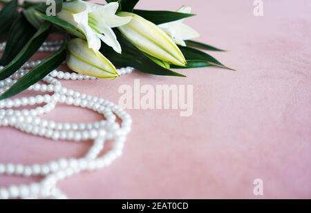 Weiße Lilien mit grünen Blättern und einer Perlenkette auf rosa Hintergrund. Sanfter Hintergrund mit Platz für Text. Stockfoto