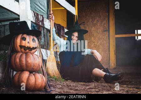 Ein Mädchen als Hexe gekleidet sitzt an einem Zaun mit einem Besen in den Händen, im Vordergrund eine gruselige Figur von Kürbissen Stockfoto