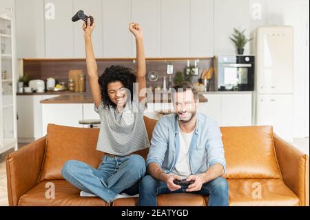 Fröhliche afrikanische Frau gewann in einem Videospiel, schreien glücklich, während und hebt die Arme, ihr Freund mit einem Joystick sitzt neben. Ein multirassisches Paar Stockfoto
