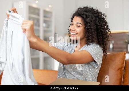 Zufriedene Biracial junge Frau mit einer Afro Frisur Auspacken Paket-Box sitzt auf dem Sofa zu Hause, aufgeregt afrikanische Frau schaut auf gelieferte Ware Stockfoto