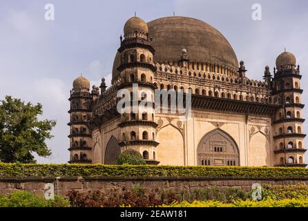 Vijayapura, Karnataka, Indien - 8. November 2013: Gol Gambaz Mausoleum. Eckansicht auf beigefarbenem Gebäude mit Türmen und geschwärzter Kuppel hinter grüner Pflanze Stockfoto