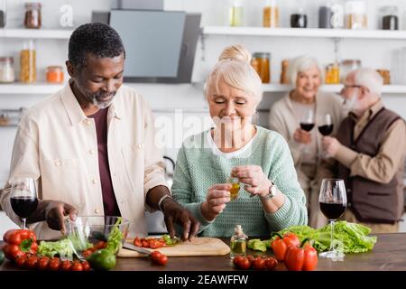 afroamerikanischer Mann und glückliche ältere Frau, die in der Nähe Salat zubereitet Pensionierte Freunde auf verschwommenem Hintergrund Stockfoto
