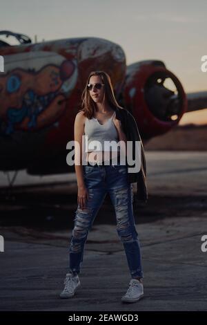 Ein großes blondes Teenager-Mädchen trägt zerrissene Jeans und ein Weiße Tank Top Posen für Porträts von verlassenen Flugzeugen bedeckt In Graffiti in der Wüste wild Stockfoto