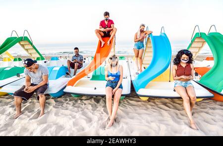 Multirassische Freunde mit geschlossener Gesichtsmaske mit Tracking-App mit Handy-Smartphone - gelangweilte junge Menschen sitzen an Strand Stockfoto