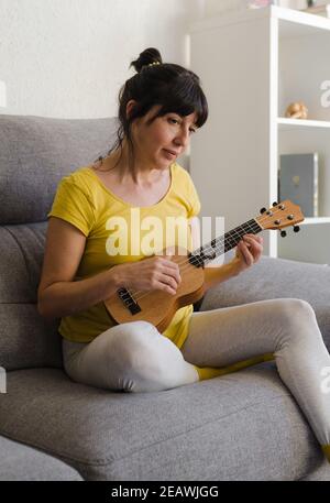 Junge Brünette Frau mit ihren Haaren gebunden zurück spielen die Ukulele auf ihrem Sofa sitzen. Sie spielt gerne ein Lied zu Hause. Stockfoto