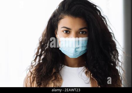 Präventionsmaßnahmen. African american hübsche Brünette Frau in Freizeitkleidung stehen drinnen mit schützenden medizinischen Maske auf ihrem Gesicht und suchen direkt Stockfoto