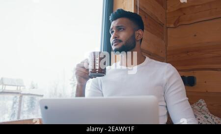 Porträt eines hübschen indischen Mannes, der Kaffee trinkt und am Laptop neben dem großen Fenster arbeitet. Hochwertige Fotos Stockfoto