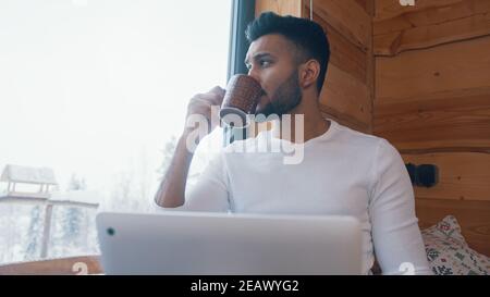 Nahaufnahme. Junger inder trinkt Kaffee am Fenster und schaut draußen auf den Schnee. Hochwertige Fotos Stockfoto