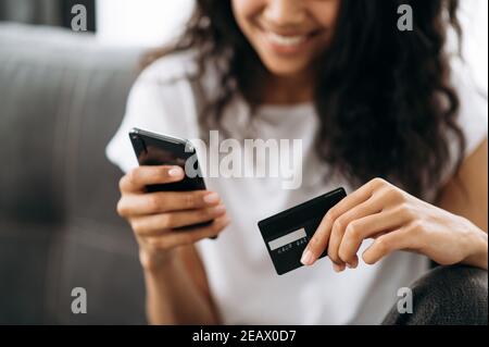 Eine Kreditkarte und ein Smartphone in weiblicher Hand. Fröhliche afroamerikanische junge Frau in Unschärfe sieht auf dem Handy-Bildschirm, Online-Shopping. Kreditkarte A Stockfoto