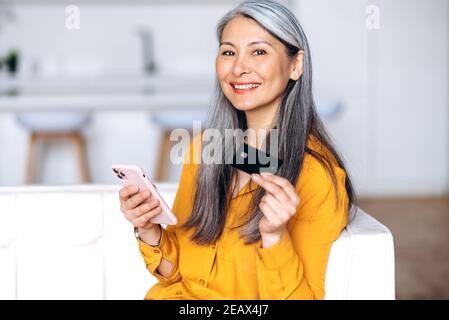 Happy mittleren Alters lächelnde grauhaarige Dame, ist online einkaufen. Fröhliche asiatische ältere Frau hält die Kreditkarte und ihr Smartphone, Online-Shopping, o Stockfoto