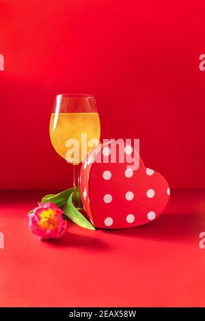Glas Champagner, herzförmige Geschenkbox und Blume auf rotem Hintergrund. Liebe romantische Feier des Valentinstag, Frauentag, Muttertag Stockfoto
