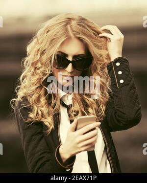 Blonde Geschäftsfrau mit Handy auf Stadt Straße stilvoll Mode-Modell mit langen lockigen Haaren in schwarzer Jacke und Sonnenbrille Stockfoto