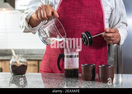 Horizontale Aufnahme eines Mannes in einer roten Schürze, der kochendes Wasser in eine französische Presse legt, um den Kaffee vorzubereiten. Barista, dessen Gesicht nicht sichtbar ist. Selektiver Fokus Stockfoto