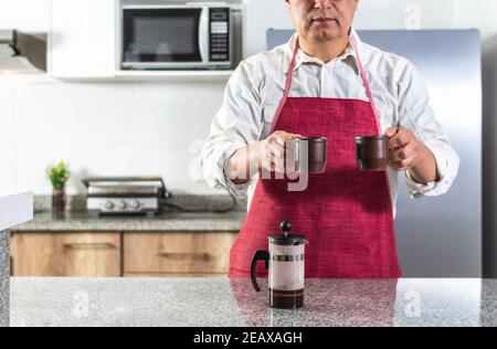 Horizontales Foto eines unsichtbaren Mannes, der eine rote Schürze trägt und zwei Tassen Kaffee auf einer Küchentheke oder Cafeteria neben einer französischen Presse anbietet. Selektiv Stockfoto