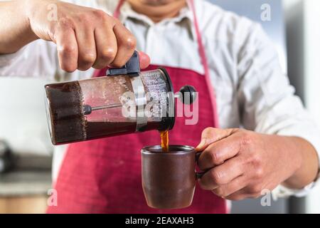 Nahaufnahme der Hände eines Mannes ein Mann mit einer roten Schürze, dessen Gesicht nicht sichtbar ist, der frischen Kaffee aus einer französischen Presse in eine Tasse gießt. Selektiver Fokus Stockfoto