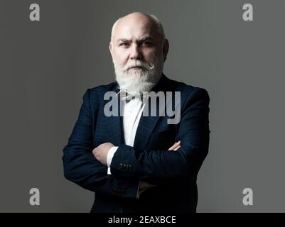 Alter Mann mit gekreuzten Armen. Grauhaarige Senior Business Mann trägt eleganten Anzug und Krawatte über grauen Hintergrund, lächelnd zuversichtlich. Stockfoto