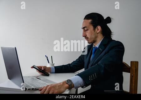 Geschäftsmann am Tag der Arbeit, Kommunikation per Smartphone während der Verwendung seines Computers, trägt einen monochromatischen blauen Anzug und eine Titanuhr Stockfoto