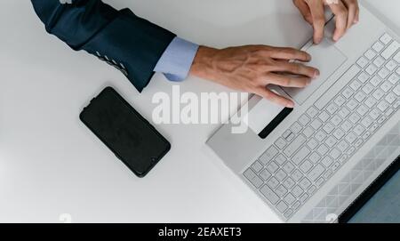 Overhead-Ansicht der Hände eines venösen Mannes in einem monochromatischen blauen Anzug Eingabe am Computer, mit einem Handy auf dem Tisch, Arbeitstag. Stockfoto