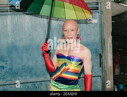 Porträt einer asiatischen queeren Person in Regenbogenfarben Kleid gekleidet, trägt rote Handschuhe und hält einen Regenbogenfarben Regenschirm in Bangkok Thailand. Stockfoto