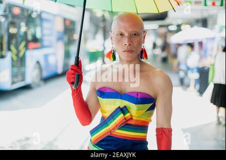 Porträt einer asiatischen queeren Person in Regenbogenfarben Kleid gekleidet, trägt rote Handschuhe und hält einen Regenbogenfarben Regenschirm in Bangkok Thailand. Stockfoto