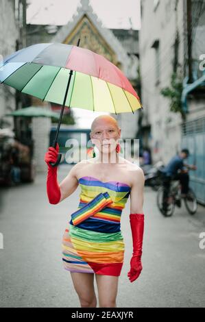 Porträt einer asiatischen queeren Person in Regenbogenfarben Kleid gekleidet, trägt rote Handschuhe und hält einen Regenbogenfarben Regenschirm in Bangkok Thailand. Stockfoto