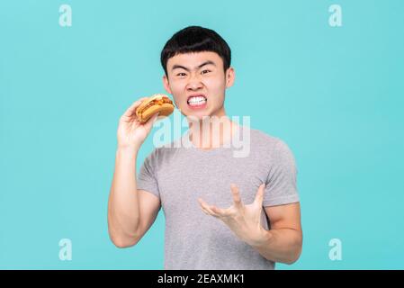 Diät hungrigen asiatischen Mann mit lustigen Gesichtsausdruck beim Halten hamburger in der Hand isoliert auf hellblauem Hintergrund