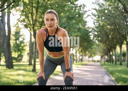 Ruhen Sie sich eine Weile aus. Schöne sportliche Frau in Sportbekleidung, die an einem sonnigen Sommertag beim Joggen in einem grünen Park eine Pause einnimmt Stockfoto