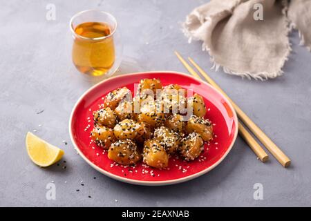 Traditionelle сhinese frittierte Äpfel in Karamell mit Sesam auf Roter Vintage-Teller mit Essstäbchen mit einem Glas Grün Tee auf grauem Strukturhintergrund Stockfoto