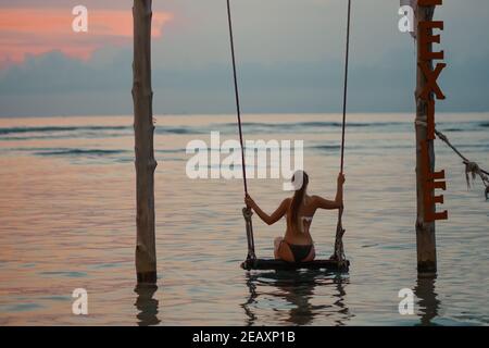 Gebräunte Frau, die auf einer Schaukel im Wasser steht. Sonnenuntergang und Meer im Hintergrund. Nahaufnahme. Relax und Urlaub Konzept. Stockfoto
