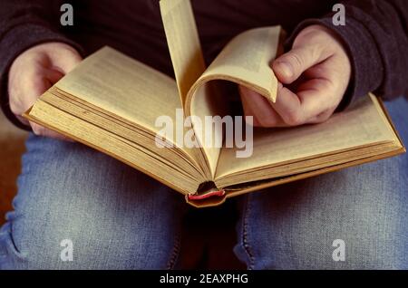 Ein Mann blättert durch ein offenes Buch, das ihm zu Füßen liegt. Das Buch ist im Hardcover. Seitenansicht, selektiver Fokus. Stockfoto