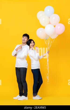 Attraktive fröhliche junge asiatische Liebhaber glücklich hält bunte Ballons auf Gelber Studiohintergrund Stockfoto