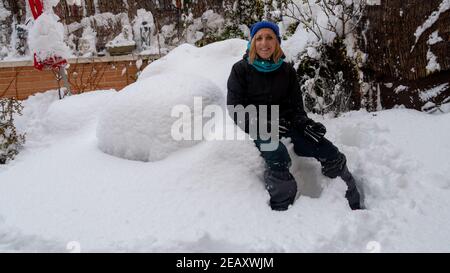Fröhliche Mitte-Alter blonde Frau in einem Mantel sitzen in der Schnee im Hinterhof im Winter Stockfoto