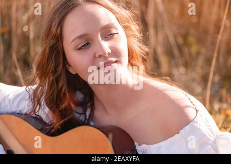 Junge hübsche Frau mit langen Haaren Musiker spielen akustische Gitarre bei Sonnenuntergang Feld im Sonnenlicht. Stockfoto