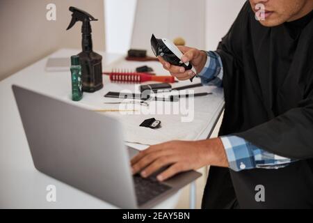 Fokussierter Mann mit verschiedenen Haarwerkzeugen auf dem Schreibtisch suchen Auf den Bildschirm von Laptop in Home-Wohnungen Stockfoto