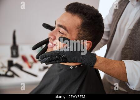 Schöner Mann ruht sich im Friseurladen aus, während der Barbier legt Kosmetisches Öl auf der Haut Gesicht Stockfoto