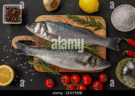 Zwei frische Seebaßfische mit Rosmarin und Gemüse auf schwarzem Tisch. Meeresfrüchtekonzept. Stockfoto