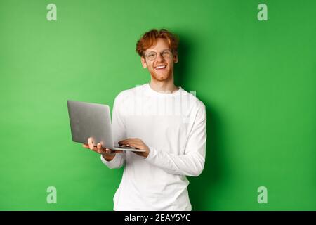 Fröhlicher Rotschopf Freiberufler in Brille lächelnd an der Kamera, arbeiten auf Laptop, während über grünem Hintergrund stehen Stockfoto