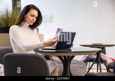 Schöne Geschäftsfrau, die am Telefon tippt, während sie mit einem Laptop am Tisch sitzt. Stockfoto