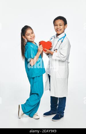 Zukünftige Herzärzte. Gruppe von multiethnischen Kindern tragen medizinische Uniformen, halten Kinder Form des Herzens in den Händen Stockfoto