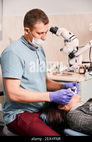 Junger Mann Zahnarzt in medizinische Maske mit zahnmedizinischen diagnostischen Mikroskop während der Behandlung von Patienten Zähne. Frau, die im Stuhl liegt, während der zahnärztlichen Kontrolle im modernen medizinischen Zentrum. Konzept der Zahnmedizin. Stockfoto