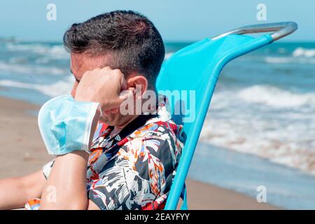 Ein junger Kaukasianer, der ein hawaiihemd trägt, sitzt in einem Liegestuhl am Strand, lehnt seinen Kopf in die Hand und trägt seine Einweg-Gesichtsmaske i Stockfoto