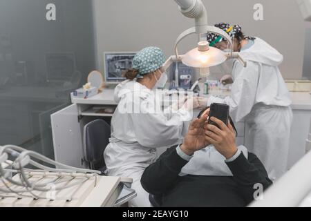 Patient mit Smartphone in der Zahnarztpraxis. Zwei Ärztinnen Vorbereitung des Verfahrens. Konzept der Zahngesundheit und Hygiene Stockfoto