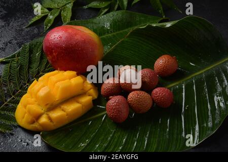 Frische Mangofrucht mit in Scheiben geschnittenen Mangostückchen auf einem Teller und frischer und geschälter Litschi, die die rote Haut und das weiße Fleisch auf dunklem Hintergrund zeigt. Stockfoto