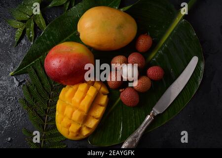 Frische Mangofrucht mit in Scheiben geschnittenen Mangostückchen auf einem Teller und frischer und geschälter Litschi, die die rote Haut und das weiße Fleisch auf dunklem Hintergrund zeigt. Stockfoto