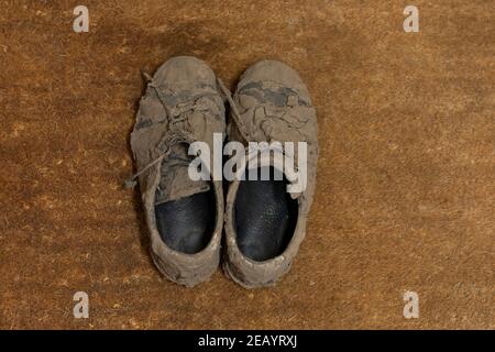 Draufsicht auf schmutzige getrocknete schlammige und unordentliche Sneaker Schuhe Völlig mit Schlamm bedeckt, der unerkennbar aussieht, während er auf einem liegt Braune Coco-Fußmatte Stockfoto